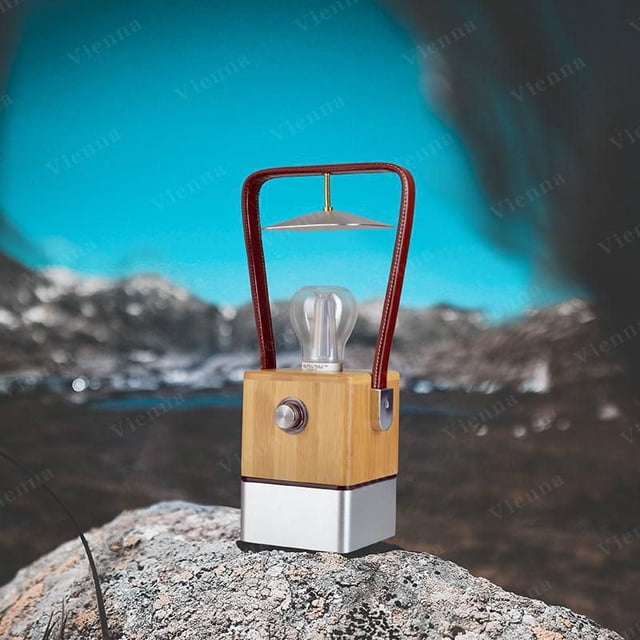 Tru De Light Vienna LED Lamp with bamboo and aluminum base, patented transparent LED bulb, and leather handle, placed on a rock with blurred mountains and blue sky, ideal for camping and outdoor use.
