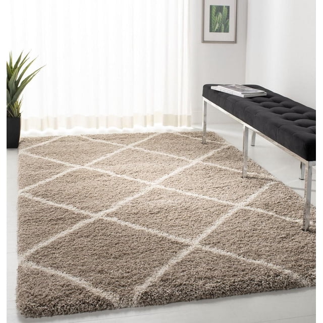 Safavieh Hudson Amias Geometric Shag Area Rug featuring a plush beige shag pile with a white geometric diamond pattern, displayed in a minimalist room with light flooring, a black tufted bench with chrome legs, a potted plant, and sheer white curtains, suitable for adding texture to modern living spaces.