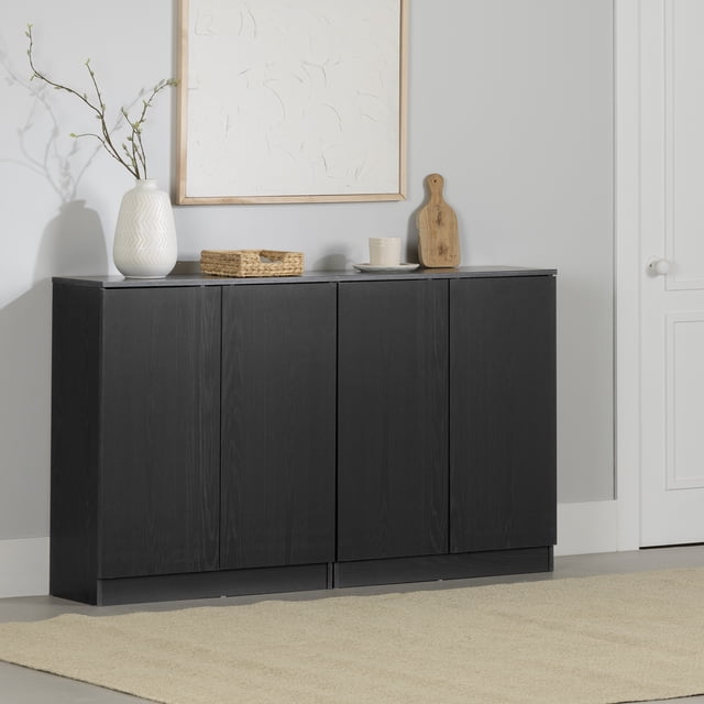 South Shore Arum Ensemble de 2 Bahuts avec 2 Portes en Chêne Noir, Montrés Côte à Côte dans une Pièce Moderne. Ces Armoires Sans Poignées Offrent une Finition Chêne Noir Mat, des Portes à Pousser pour Ouvrir et Repose sur une base de podium. les Articles Décoratifs sur le Dessus Incluent un vase Blanc avec des branches, un Panier Tressé, une Tasse sur une Soucoupe et une Planche à Découper en Bois. Positionnés contre un Mur Gris Pâle avec une Oeuvre Texturée Blanche, les Bahuts Offrent un Rangement de Cuisine Modulaire avec des Tablettes Ajustables,