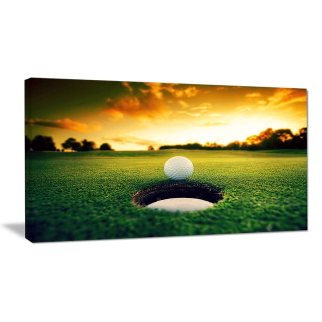 Design Art Golf Ball near Hole Landscape Artwork Canvas Print featuring a close-up of a white golf ball resting on the edge of a golf hole on green turf, under a vibrant sunset sky with distant tree silhouettes, gallery wrapped on a 1-inch wood subframe, ready to hang as wall art.