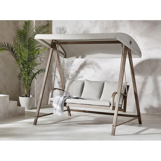 Better Homes & Gardens Prado 3-Seat Steel Canopy Outdoor Porch Swing with Cushions, Brown, featuring a rust-resistant powder-coated steel frame with faux wood finish, light beige adjustable canopy and cushions, three back cushions, and a light throw blanket, shown indoors near a potted palm and concrete stairs, suitable for patio furniture relaxation.