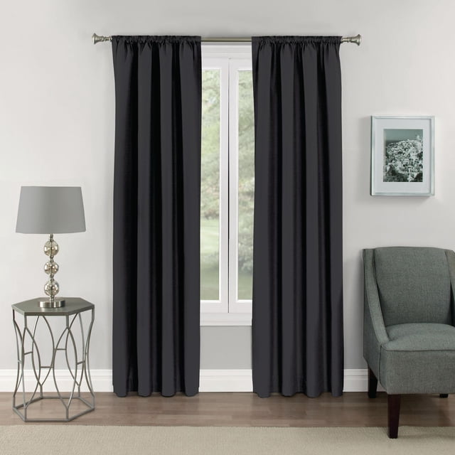 Eclipse Samara 63" rod pocket blackout panel in dark charcoal hanging on a silver curtain rod over a white-framed window in a living room with a grey upholstered chair, geometric metal side table with silver lamp, and framed black-and-white photo. This noise-reducing, energy-saving curtain blocks over 99% of light and reduces drafts, ideal for improving sleep quality.