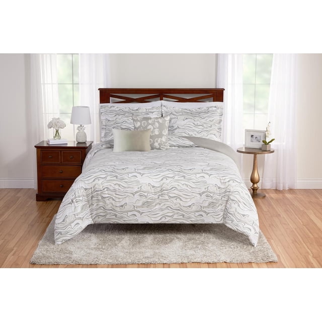 Marble 5 Piece Comforter Set on a bed with a wooden headboard in a bright room. The reversible comforter and shams feature grey marble swirl patterns on a creamy background in soft faux suede polyester. The set includes three decorative pillows in solid beige and complementary prints, plus an adjustable bed skirt. It is machine-washable and styled with a wooden nightstand holding a white lamp and flowers, and a round side table with a framed photo and plant.