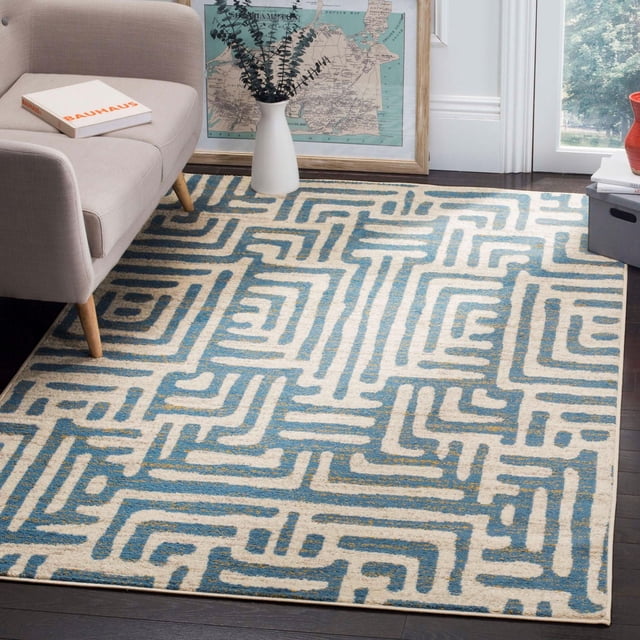SAFAVIEH Amsterdam Elvan Geometric Area Rug with a blue and cream geometric tribal pattern on a soft, durable synthetic pile, placed on dark hardwood flooring beside a beige mid-century modern sofa and a white vase with eucalyptus branches, with a framed vintage map leaning against a white wall, ideal for adding rustic-chic décor to living rooms.