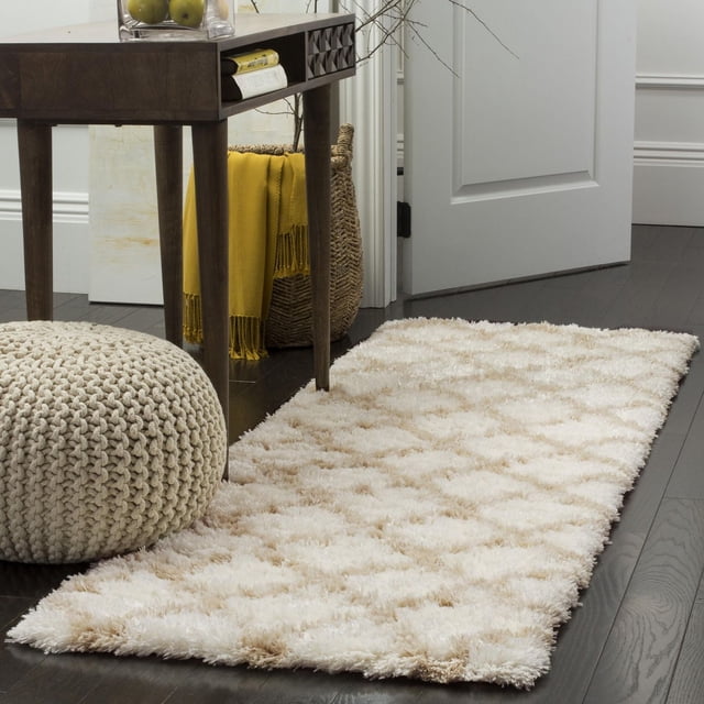 Safavieh Indie Careen Geometric Shag Area Rug with a plush cream and beige geometric pattern and thick textured pile, power-loomed from a blend of yarns and fibers, placed on dark hardwood flooring beside a dark wood console table, knitted pouf, and woven basket with yellow throw, suitable for adding warmth and texture to home spaces.