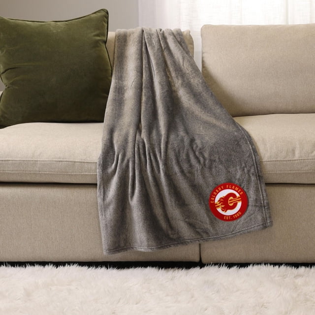 NHL, NBA, MLB Grey Sports Throw Blanket, 50" x 60", draped over a beige sofa with a green cushion, featuring a red Calgary Flames logo patch, made of soft plush polyester for warmth and comfort, suitable for indoor and outdoor use.