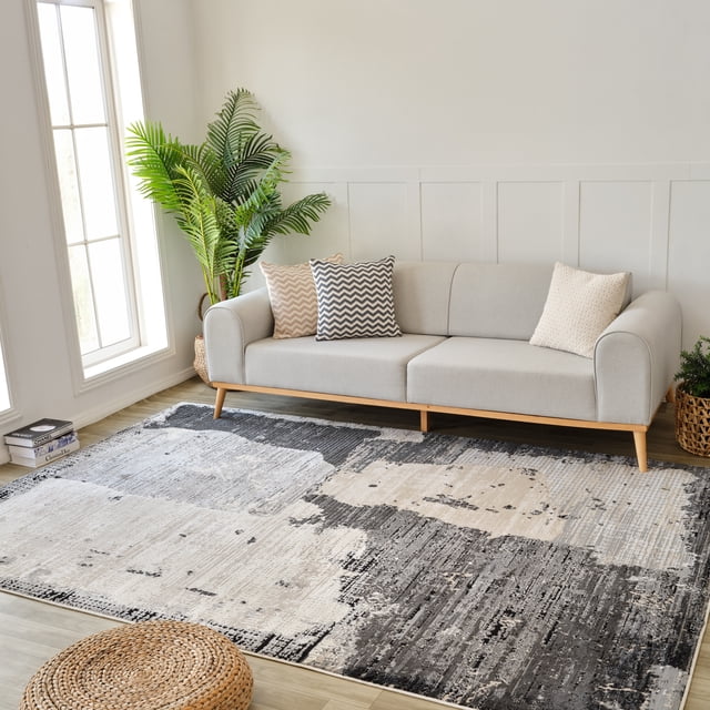 Rug Branch Nomad Abstract Indoor Area Rug in black and beige with a modern abstract pattern, measuring 7'10" x 10'10", displayed in a bright living room with a light gray sofa with wooden legs and three patterned pillows, a tall green potted plant near a large window, and a woven round ottoman on light wood flooring, ideal for living room, bedroom, dining room, or kitchen.