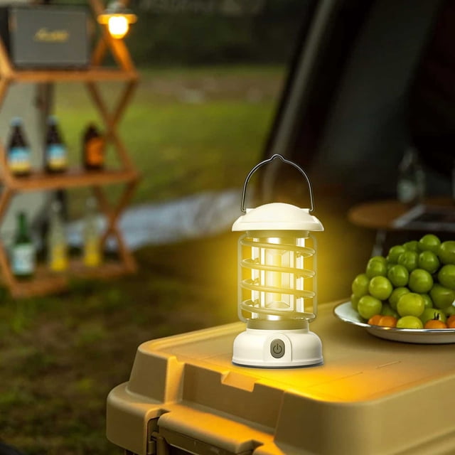 SuoKom Camping Lantern Portable Retro Camping Lamp USB Rechargeable Camping Lantern with a white plastic body, dimmable LED light, and a hanging hook. The lantern is shown illuminated on a camping table with a plate of green fruits nearby, set against a blurred outdoor background. Ideal for outdoor camping, hiking, and emergencies.