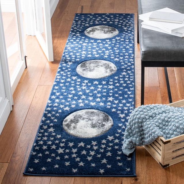Safavieh Carousel Kids Tapis Lune dans les Étoiles Est un Coureur Rectangulaire Bleu Présentant Trois phases de Lune Grises et Blanches Entourées d'Étoiles Blanches Dispersées, Tissé Mécaniquement avec des Fils Synthétiques Durables pour les Chambres d'Enfants, Placé sur un Plancher de Bois Franc à Côté d'Un Banc Gris et d'Une Caisse en Bois avec une Couverture Tricotée Bleue.