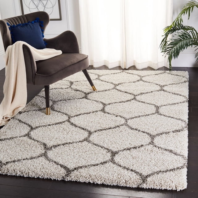 Safavieh Hudson Tapis Shag Géométrique Arline en Ivoire avec une Épaisse Peluche Shag à Poils Longs Présentant des Motifs de Carreaux en Forme d'Oignon Gris Foncé, Installé sur un Plancher de Bois Franc Foncé à Côté d'Une Chaise en Velours Gris Foncé avec des Pattes à Pointe Dorée, un Coussin à Pompons Bleus et un Jeté en Tricot Crème dans une Pièce Lumineuse avec des Rideaux Transparents et une Plante Verte, Idéal pour Ajouter de la texture aux Espaces de Vie Modernes.