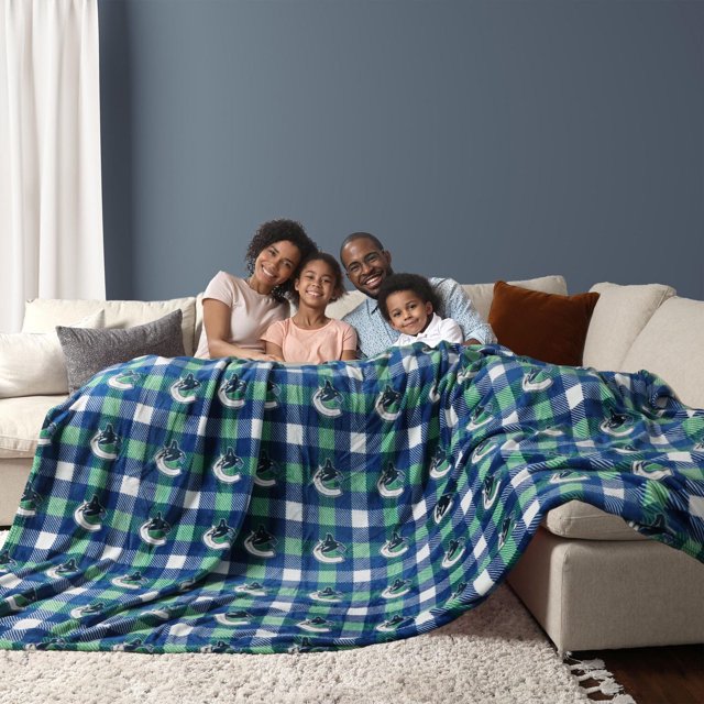 NHL Vancouver Canucks Game Day Blanket, 60" x 96", draped over a beige sectional sofa with a family of four sitting behind it, featuring a blue, green, and white plaid pattern with repeated Vancouver Canucks logos, set in a living room with a blue accent wall and neutral-toned pillows, suitable for cozying up during game nights or movie evenings.