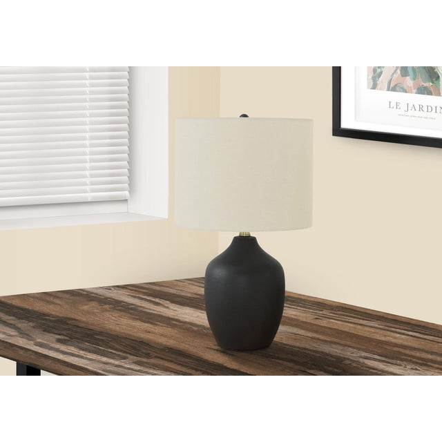 Monarch Specialties Lighting 22-inch transitional style table lamp with a smooth black ceramic base and ivory linen drum shade, topped with a round black finial, featuring a 3-way switch, placed on a dark wood grain side table near a window with white blinds and framed wall art.