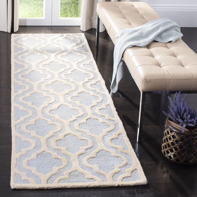 Safavieh Cambridge Kirsten Geometric Area Rug with a light grey background and cream raised trellis pattern, hand-tufted wool texture, displayed on dark hardwood flooring next to a beige tufted bench with chrome legs and a light blue knitted throw, accompanied by a decorative pot of purple flowers.