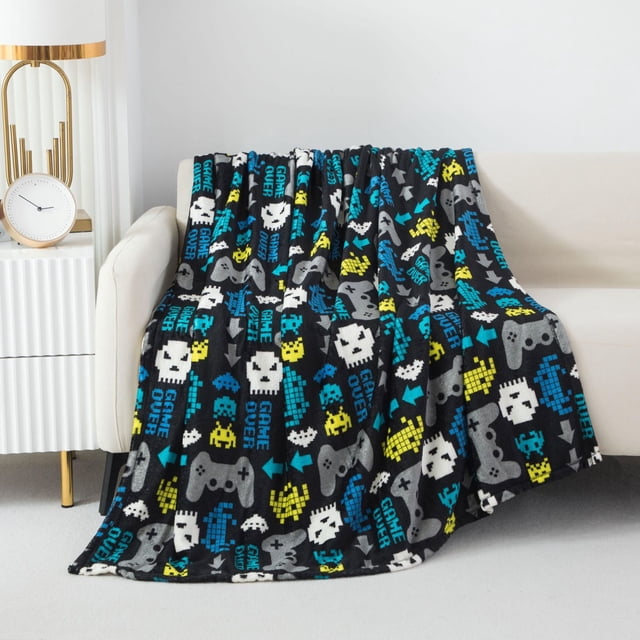 Mainstays Kids Couverture Jetée en Peluche Game Over avec Fond Noir et Icônes de Jeux Vidéo Pixélisées en Bleu, Jaune, Blanc et Gris Incluant des Manettes de Jeu et un Texte « Game Over », Fabriquée en polyester Doux 100 % Mesurant 50 par 60 Pouces, Drapée sur un sofa beige, Idéale pour Ajouter Chaleur et style aux Chambres d'Enfants et Adaptée au Lavage Facile à la machine.