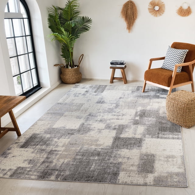 Rug Branch Jules Abstract Indoor Area Rug in grey beige with a modern abstract geometric pattern, placed on light wood flooring in a bright room featuring a large black-framed arched window, a rust-coloured armchair with a chevron cushion, a woven basket planter with green foliage, a small wooden stool with books, and natural fibre wall decor.