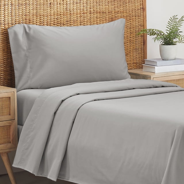 California Design Den 100% Organic Cotton Sheet Sets in a soft grey colour, featuring eco-friendly percale weave for breathability and cooling. The twin-size set includes a flat sheet, elasticized fitted sheet with deep pockets, and a standard pillowcase. Displayed on a neatly made bed with a rattan headboard and a bedside table holding books and a small plant.