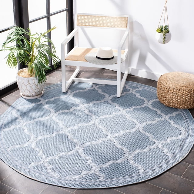 Safavieh Bermuda Corie Geometric Quatrefoil Outdoor Area Rug in round shape with light blue base and white quatrefoil pattern, placed on dark tile floor with a white wooden chair with woven seat and backrest holding a straw hat, a woven round ottoman, potted green plant, and hanging succulent planter in a sunlit space, suitable for outdoor patios or decks.