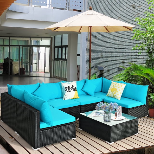 Patiojoy 7-Piece Patio PE Rattan Sectional Sofa Furniture Set in turquoise, featuring a modular design with 4 armless sofas, 2 corner sofas, and a coffee table with a tempered glass top. The set is crafted with weather-resistant black PE wicker and powder-coated steel for durability. Thick turquoise cushions with zippers provide comfort and are easy to clean. The setup is displayed on a wooden deck with a beige umbrella and decorative pillows, surrounded by greenery.