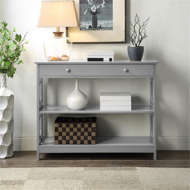 Convenience Concepts Omega Storage Console Table in Gray Wood Finish, featuring a modern geometric side design, a single drawer for concealed storage, and two open shelves for display. Made of MDF with a sleek grey finish, it measures 31.5 inches high, 39.5 inches wide, and 12 inches deep. Shown in a living room setting with decorative items and a lamp on top.