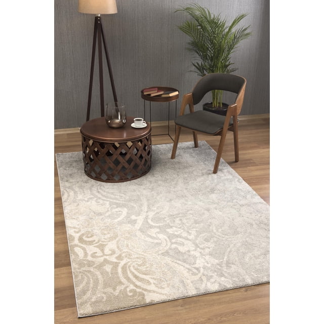 Rug Branch New Zealand Plush Collection High-Density Modern Damask Soft Durable Area Rug in beige and light grey tones with intricate vintage damask pattern, placed on hardwood floor in a modern living room setting with a dark wood frame chair upholstered in black fabric, round metal lattice coffee table with glass vase and coffee cup, small round side table with books, tripod floor lamp, and green potted plant against a textured grey wall. This high-density polyester rug is ideal for living rooms, bedrooms, or home offices.