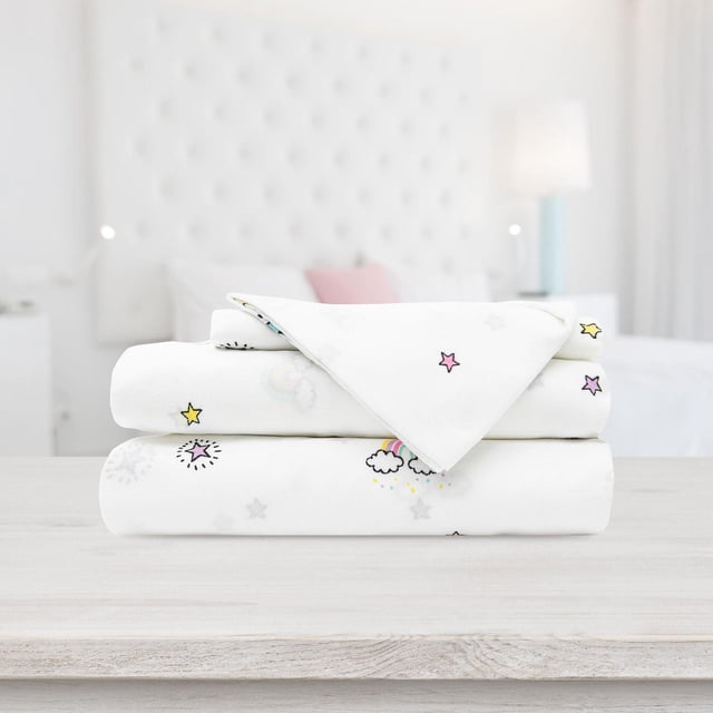 Safdie & Co. Premium Ultra Soft Bedding Printed Sheet Set 4PC Double Rainbow with folded white sheets featuring pastel stars and rainbow cloud prints, hypoallergenic and wrinkle-resistant fabric, displayed on a light wood surface in a softly lit bedroom with a tufted white headboard.