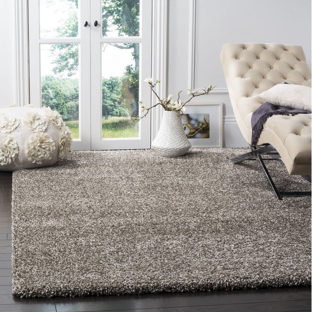 Safavieh Milan Harlow Tapis Shag Uni en Gris, Posé sur des Planchers de Bois Franc Sombres dans une Pièce Lumineuse. Ce Tapis d'Accent Moelleux Présente une pile Shag Synthétique dense pour un Entretien Facile. Il Est Habillé d'Un Fauteuil lounge beige Capitonné, d'Un vase Texturé Blanc avec des branches Florissantes, et d'Un pouf floral Blanc Décoratif Près de Portes en Verre Surplombant la Verdure.