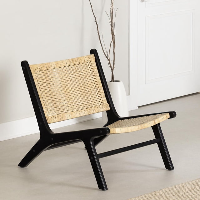South Shore Balka Rattan Lounge Chair with black teak wood frame and natural rattan woven seat and backrest, placed indoors on a light floor near a white vase with bare branches and a white wall, suitable for a boho-chic living room or bedroom relaxation area.