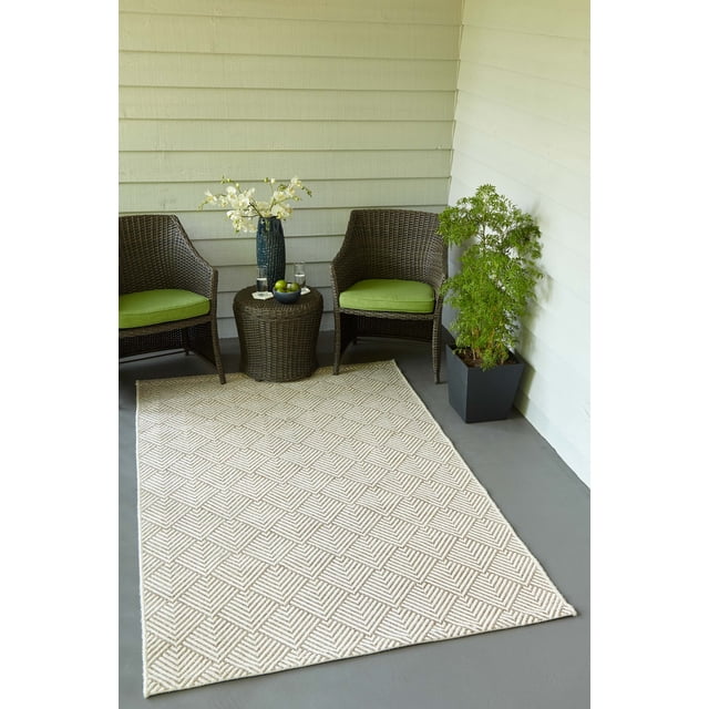 ECARPET Laura Contemporary Area Rug with a repeating geometric pattern of overlapping pointed angles in natural beige and off-white tones, made from stain-resistant polypropylene fibers. It is placed on a gray floor between two brown wicker chairs with green cushions, a round wicker side table holding a blue vase with white flowers and a bowl of green apples, and a potted green plant against light beige siding walls, suitable for both indoor and outdoor spaces.