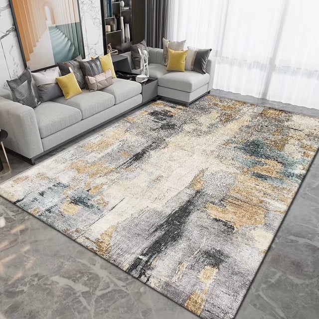 Contemporary Boho Rugs Splash Ink Abstract Printed Carpet in a large area rug size, displayed in a modern living room with grey sofas and yellow accent pillows. The rug features a grey base with bold splashes of gold, beige, and blue, creating an abstract, artistic design. Made with polyester, it offers plush softness, stain resistance, and durability, suitable for high-traffic areas and kid-friendly spaces.