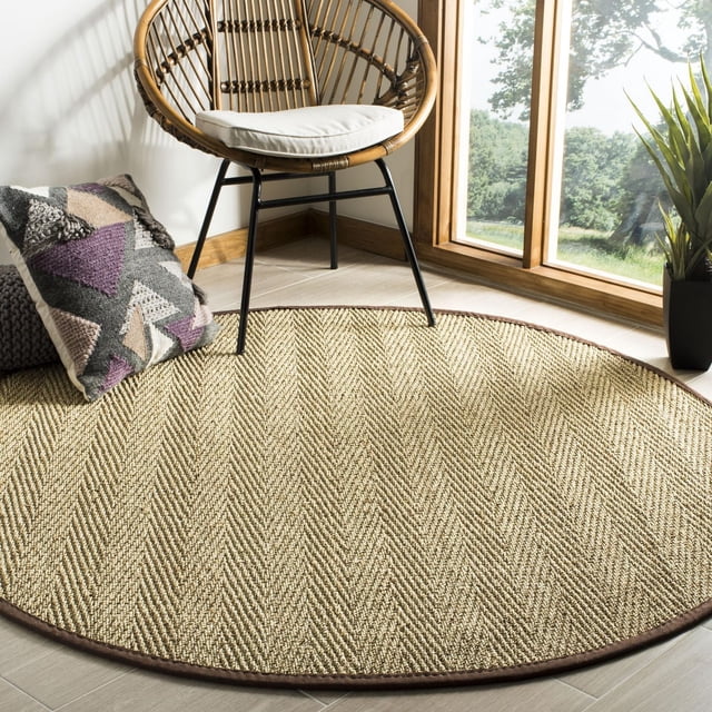 Safavieh Natural Fiber Maisy Border Area Rug, Natural / Grey, 8' X 10', a round natural fibre rug with a herringbone weave and dark brown border on light wood flooring, beside a rattan chair with a white cushion, a geometric pillow, and a plant near a window showing outdoor greenery, suitable for casual coastal or city living spaces.