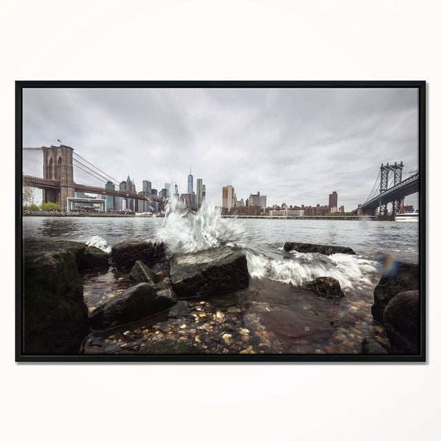 Design Art Skyline with Brooklyn Manhattan Bridges Framed Canvas Art Print showing a front-facing view of New York City skyline with Brooklyn Bridge on the left and Manhattan Bridge on the right under a grey sky, waves splashing on dark rocks in the foreground, printed on gallery-wrapped canvas with a black floating frame, ready to hang with included kit.