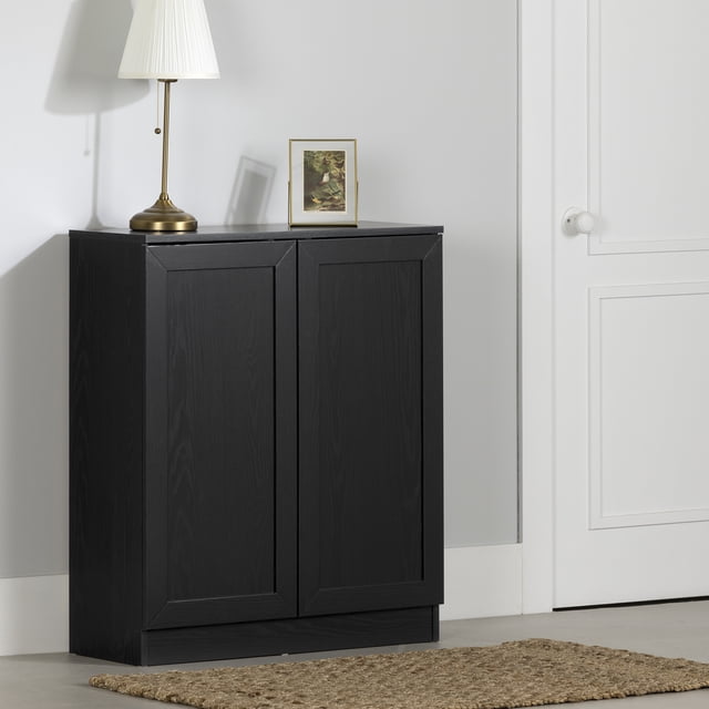 South Shore Essence Armoire de Rangement à 2 Portes en Chêne Noir avec Portes de Style Shaker et base podium, Présentée dans une Entrée avec une Lampe en Laiton et une Oeuvre Encadrée sur le Dessus, à Côté d'Une Porte Blanche et d'Un Tapis en Fibre Naturelle, Convient pour Organiser les Articles Ménagers.