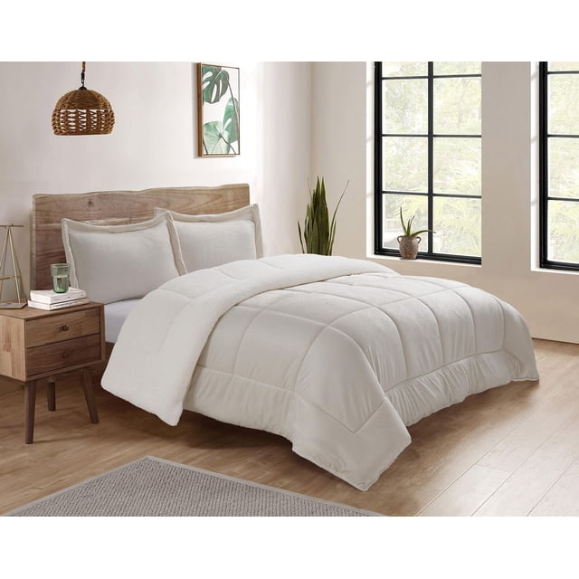 Reversible Mink & Berber Plush Sherpa Comforter Set on a bed with a natural wood headboard in a bright bedroom. The white comforter features a quilted plush side with the Sherpa fleece side folded back, paired with matching pillow shams. The room includes light wood flooring, a wooden nightstand with books and a glass, a woven pendant lamp, green plants, and large black-framed windows. This set offers reversible softness and warmth, suitable for cozy bedding and enhancing bedroom comfort.