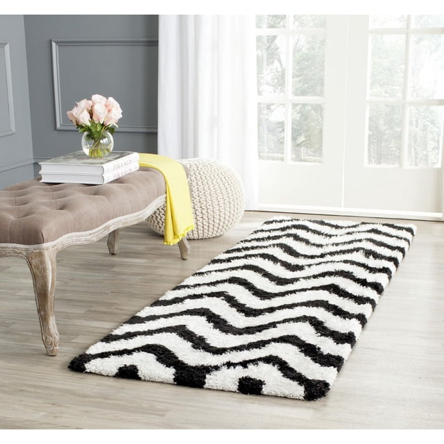 Safavieh Barcelona Carmella Zigzag Stripes Shag Area Rug with black and white zigzag stripes and plush shag texture on light hardwood floor in a bright room featuring a beige tufted bench with carved legs, yellow throw, books, pink roses in a glass vase, and a cream knitted pouf near white French doors, suitable for Mediterranean-inspired mod décor.