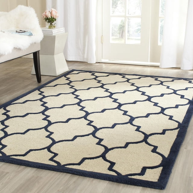 Safavieh Cambridge Kevin Geometric Area Rug with navy blue geometric trellis pattern on cream hand-tufted wool, placed on light wood floor in a bright room with white curtains and beige chair with white fur throw, ideal for modern home decor.