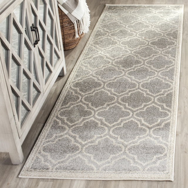 Safavieh Amherst Wilson Geometric Area Rug with a light grey and cream trellis pattern and soft cut pile, placed on a light wood floor next to a grey cabinet with glass-paneled doors and a woven basket with a fringed throw, ideal for high-traffic indoor or outdoor spaces.