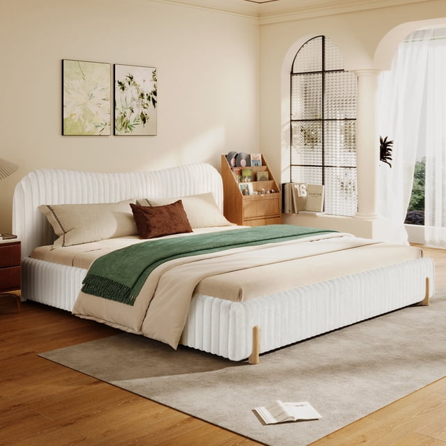 Noble House Modern minimalist upholstered bed, King size, white, featuring a curved headboard and ribbed long-pile plush fabric upholstery on a low-profile platform frame with natural rubberwood legs. The bed supports a 1.9m x 2m mattress and is set on a beige rug over wooden flooring in a bright bedroom with neutral walls, two abstract floral paintings, a wooden magazine rack, and sheer curtains on a large window.