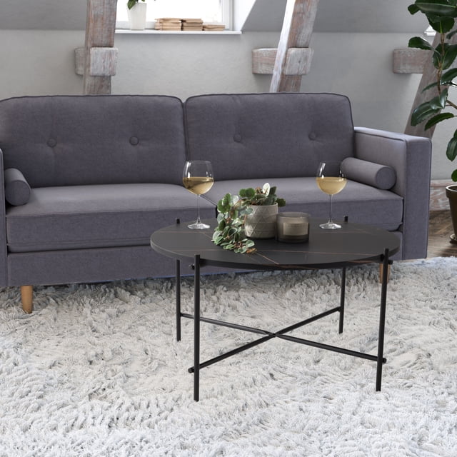 CorLiving Ayla Black Marbled Effect Engineered Wood Indoor Coffee Table with Iron Legs with a round black marbled tabletop and black powder-coated iron legs with crossbar support, placed on a light grey shag rug in front of a grey button-tufted sofa, holding two wine glasses, a potted plant, and a candle, suitable for small living rooms and modern interiors.