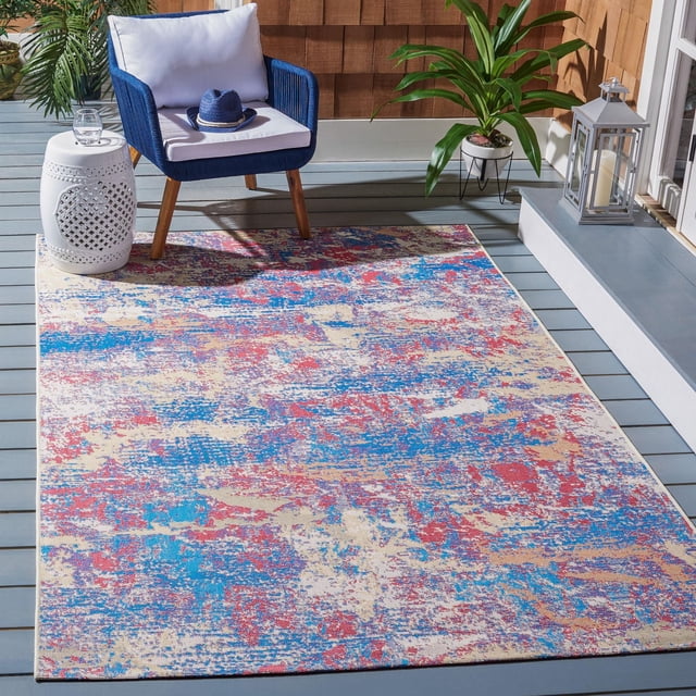 Safavieh Summer Portmont Abstract Outdoor Area Rug with a multicolor abstract pattern in blue, red, beige, and tan tones on a gray wooden deck. The durable synthetic rug resists wear, weather, staining, and fading, ideal for patios and porches. It is styled with a blue woven armchair with white cushions, a white ceramic side table holding a glass of water, a potted green plant, and a silver lantern near a sliding door, suitable for enhancing leisure outdoor spaces.