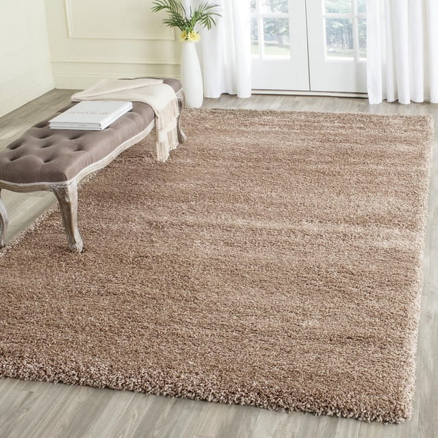 Safavieh Milan Harlow Tapis Shag Uni en Brun Doux avec Poils Shag Denses sur Plancher de Bois Clair. Ce Tapis d'Accent Rectangulaire Présente des Fils Synthétiques Faciles d'Entretien et Ajoute Chaleur et texture à la Déco Contemporaine, avec un Banc Capitonné, un Jeté beige et un vase avec de la Verdure.
