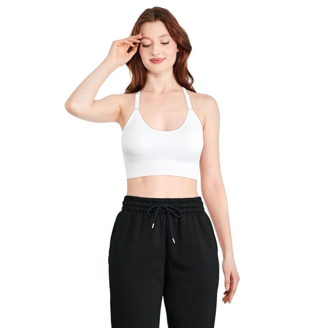 Athletic Works Women's Cami Yoga Bra in white, worn by a front-facing model with black drawstring pants. The seamless jacquard knit bra features a round neckline, adjustable straps, and Y-back design with DRI-MORE moisture-wicking fabric for comfort during workouts. It is also suitable for low-impact activities like pilates.