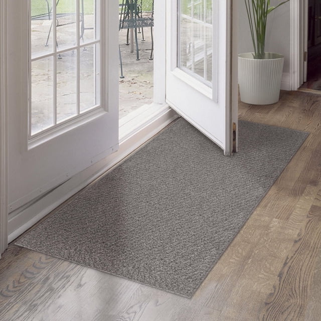 Tapis Lincoln Gris 2 Pi x 5 Pi avec Fil de Polypropylène Tufté à la Machine Se Tient sur un Plancher de Bois à l'Intérieur d'Une Entrée, Présentant une texture Grise Douce Qui Résiste aux Taches et Protège les Planchers de la Saleté, du Sel et des Débris, à Côté d'Une Ouverture de Porte Blanche pour un patio et d'Un Cache-Pot Blanc avec une Grande Plante Verte. Sa Taille le Rend Aussi Adapté à l'Utilisation comme Petit Protecteur de Plancher Intérieur dans les Entrées Ou les Couloirs.