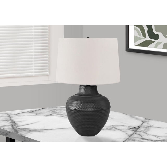 Monarch Specialties Lighting 26"h table lamp with a black metal urn-shaped base featuring a textured hammered finish and an ivory linen fabric shade with a round black finial, placed on a white marble table in a modern room with gray walls, white blinds, and framed abstract art.
