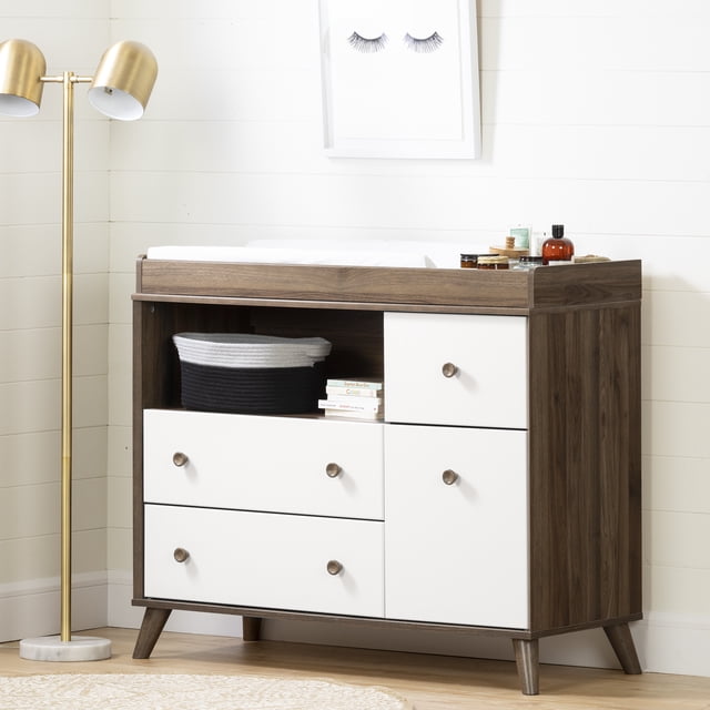 South Shore Yodi Changing Table with Drawers and Open Storage in natural walnut and white, featuring a removable top with raised edges, four drawers with wood cone-shaped handles on metal slides, and an open shelf holding a black and white woven basket and children's books, set against a white shiplap wall with a gold floor lamp and framed eyelash artwork.