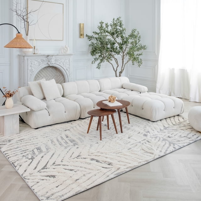 Rug Branch Nimbus Floral Indoor Area Rug in cream grey featuring a modern floral leaf pattern, placed on light hardwood flooring in a bright living room with a tufted cream sectional sofa, two nested wooden coffee tables, a white ornate fireplace, and a tall green indoor plant.