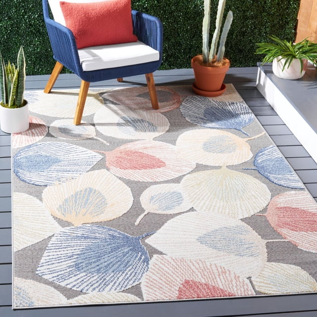 SAFAVIEH Sarasota Nash Floral Indoor/Outdoor Area Rug on a grey wooden deck, featuring a large floral pattern in muted blue, cream, and coral hues. Crafted with weather-resistant synthetic yarns, this durable outdoor rug pairs with a navy blue woven chair and coral cushion, surrounded by potted plants, ideal for enhancing outdoor seating areas.