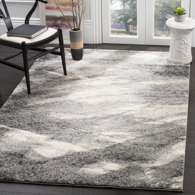 Safavieh Retro Panit Abstract Area Rug featuring a low pile polypropylene construction with a mid-century modern abstract pattern in grey, cream, and black tones, displayed on dark hardwood flooring in a bright room with a black wooden chair, a gradient ceramic vase with branches, and a white ceramic side table with a small succulent, ideal for accenting contemporary living spaces.