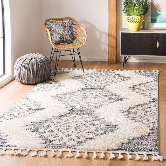 Safavieh Moroccan Tassel Shag Desmond Southwestern area rug featuring a cream and gray geometric pattern with tassels on edges, styled in a modern room with wicker chair, knitted pouf, and plant.