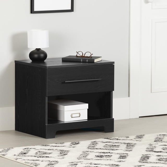 South Shore Primo 1-Drawer Nightstand for Adult in black oak finish featuring a modern rectangular design with a single drawer and metal handle above an open storage space holding a white box. The flat top holds a black lamp with white shade and a book with glasses, set against a light grey wall and white door on a light floor with a patterned rug. Its compact size is also suitable for bedside storage of personal items.