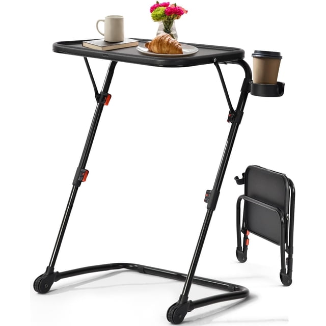 Homy Casa Folding Accent Table in black with adjustable height legs, featuring a rectangular scratch-resistant plastic surface holding a croissant on a white plate, a beige mug, and a closed book. It includes a fixed cup holder on the right side with a disposable coffee cup. The matte black metal frame has non-slip feet and locking clips on the legs. A folded version is shown beside it, demonstrating its space-saving foldable design suitable for small apartments or dorm rooms. The table is also often used as a portable laptop desk for flexible workspaces.